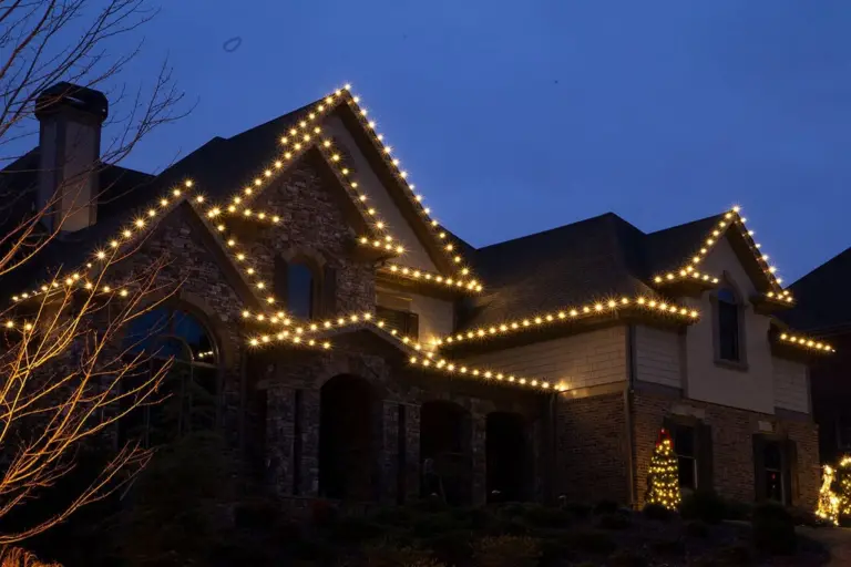 Roofline Christmas lights on a two-story stone home at dusk with a lit tree in the front yard