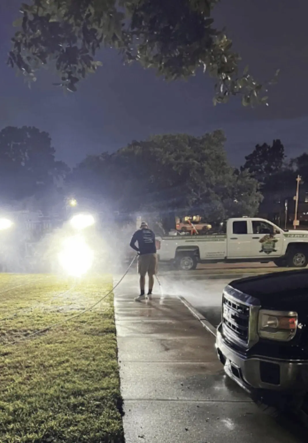 A crew member washing a sidewalk at night under work lights beside a Jade service truck
