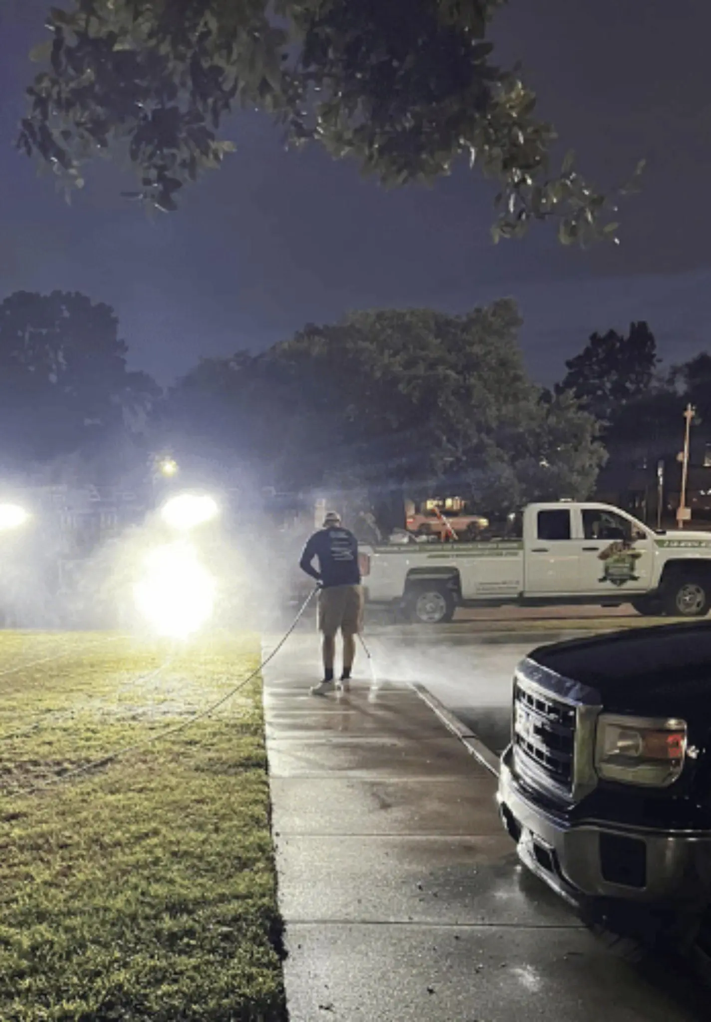 A crew member washing a sidewalk at night under work lights beside a Jade service truck