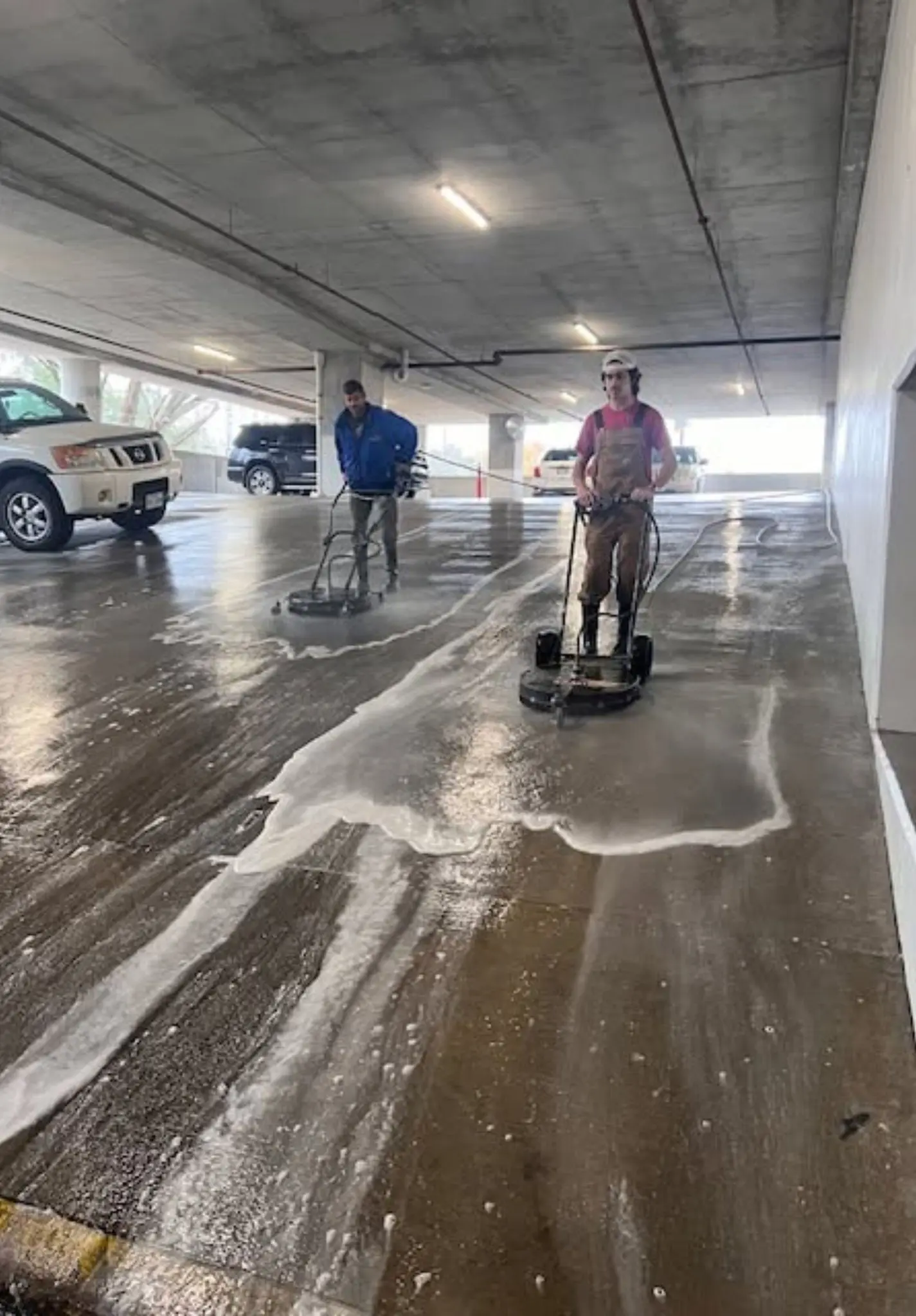 Two crew members running surface cleaners across a wet parking garage deck