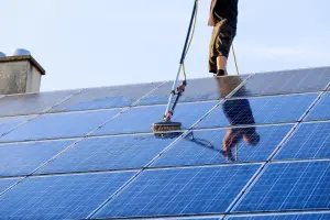 A worker on a roof scrubbing solar panels with a rotary brush on a pole