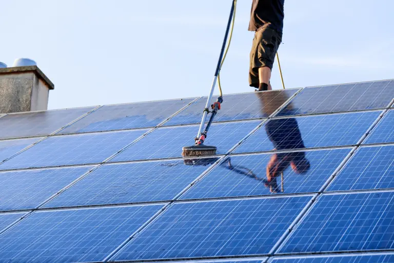 A worker on a roof scrubbing solar panels with a rotary brush on a pole