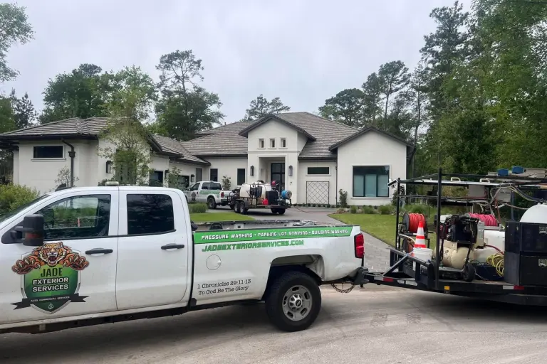 A Jade truck and equipment trailer parked in the driveway of a large white home surrounded by pines