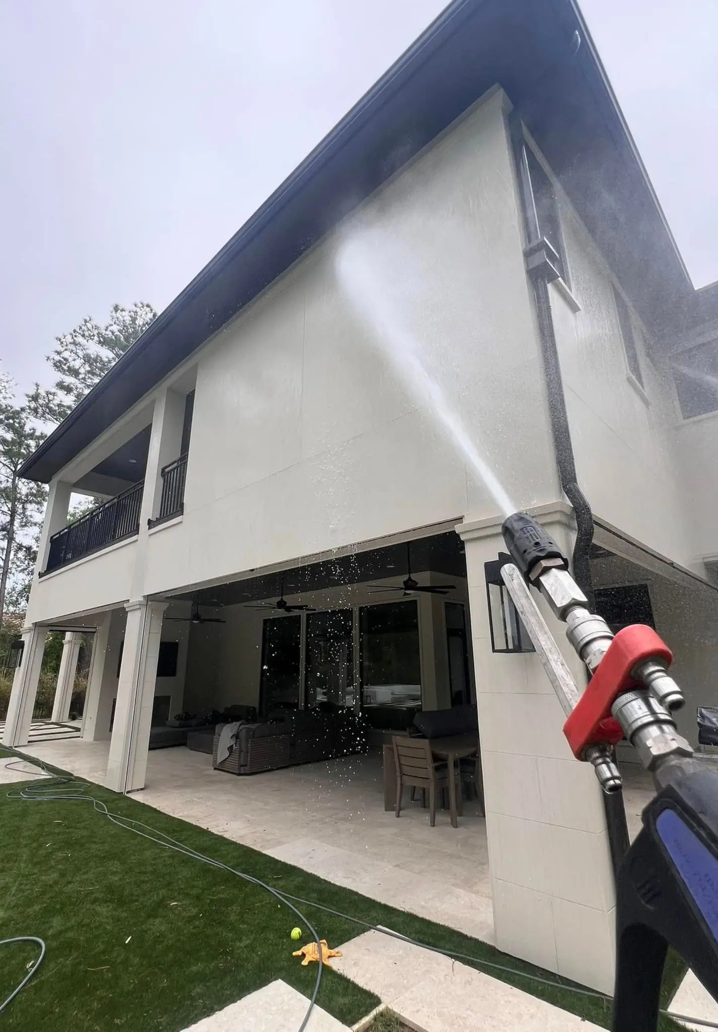 A soft wash wand rinsing the upper wall of a white two-story home above a covered patio