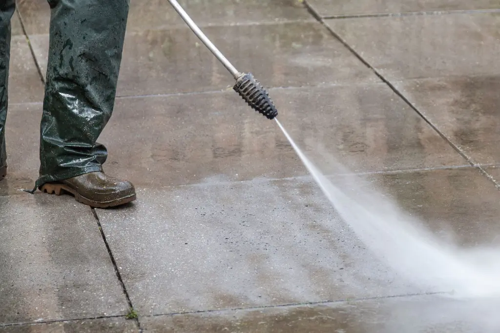 A worker in boots washing a concrete sidewalk with a pressure washer wand