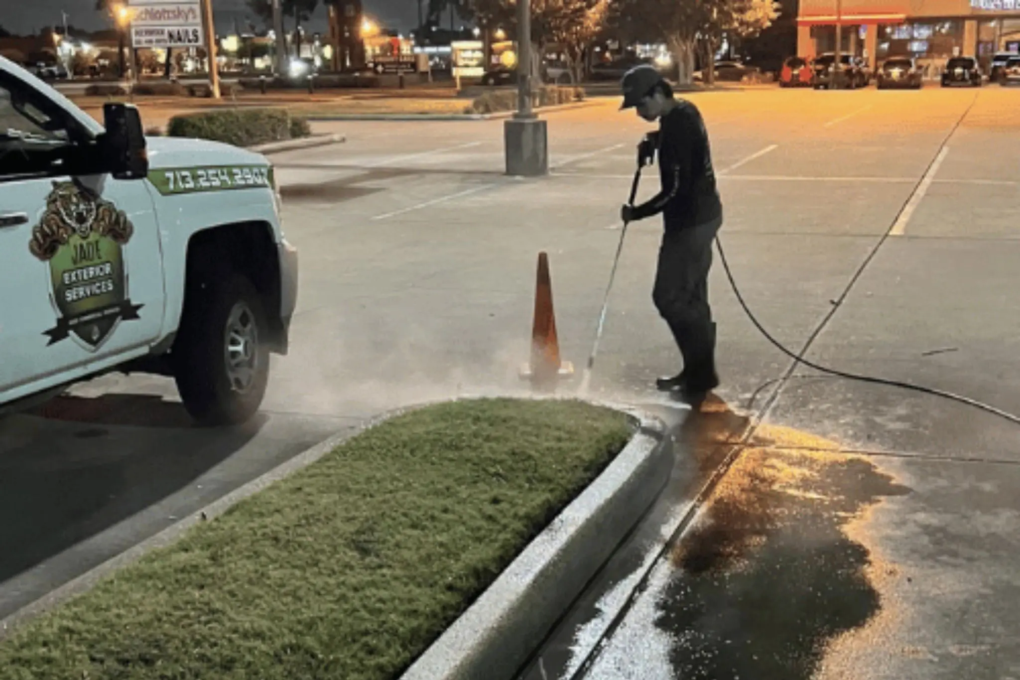 A crew member pressure washing the curb line of a commercial parking lot next to a traffic cone