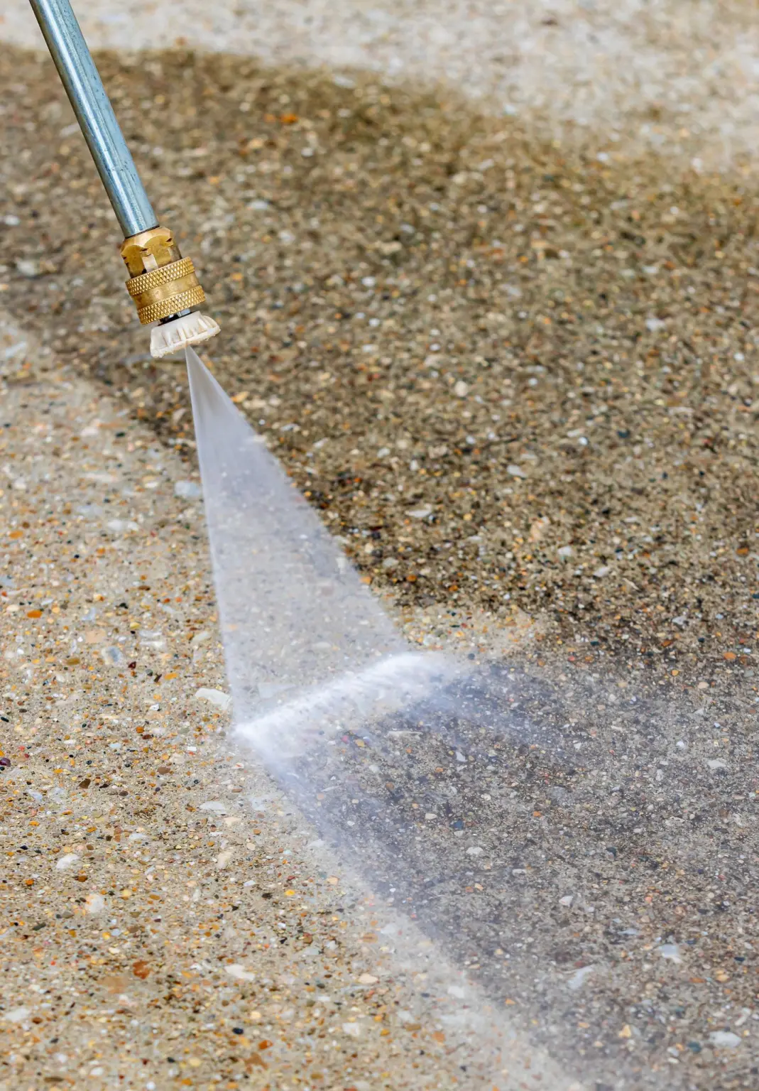A pressure washer wand cutting a clean stripe across dirty exposed aggregate concrete