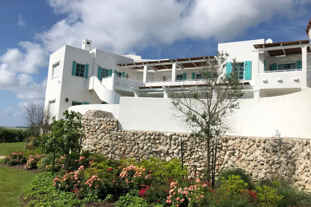 A white multi-level home with teal shutters above a stone retaining wall and rose beds