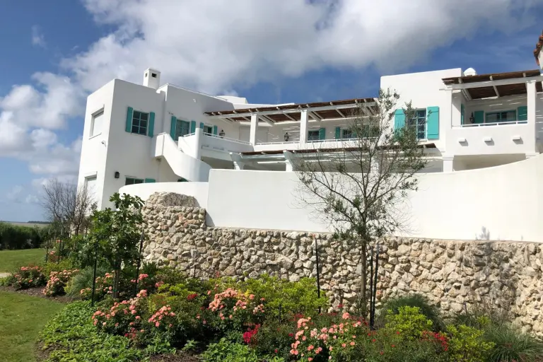 A white multi-level home with teal shutters above a stone retaining wall and rose beds